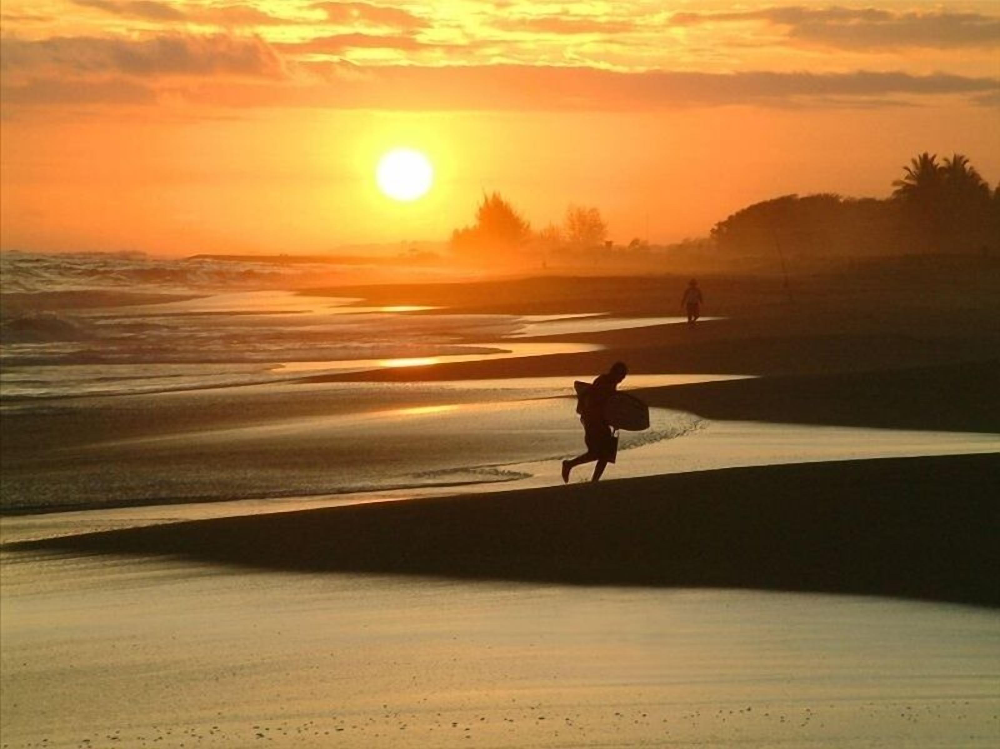 Escena de playa con un atardecer, olas y dos personas caminando.
