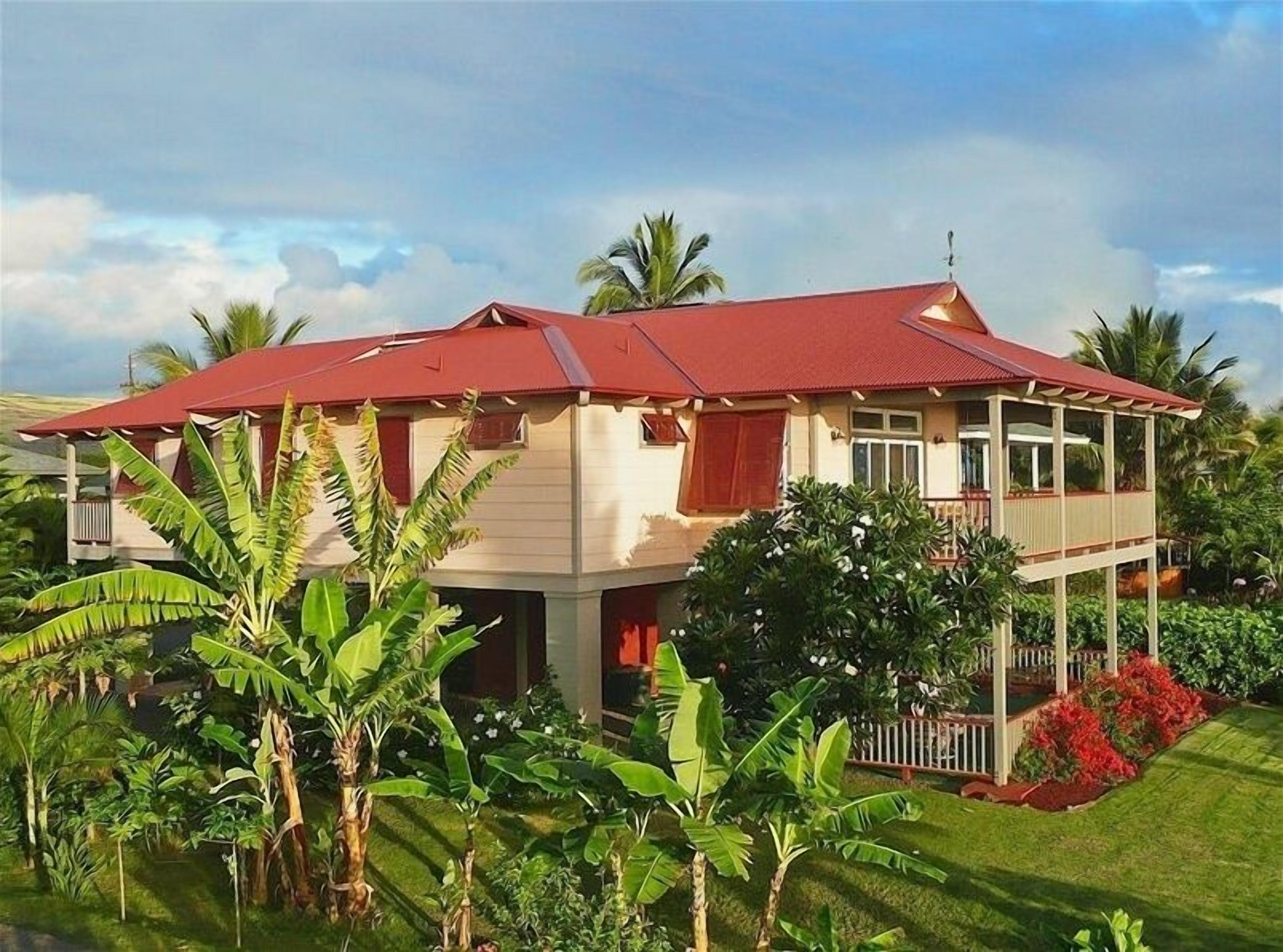 A two-story house with a red roof, surrounded by lush greenery and banana plants.