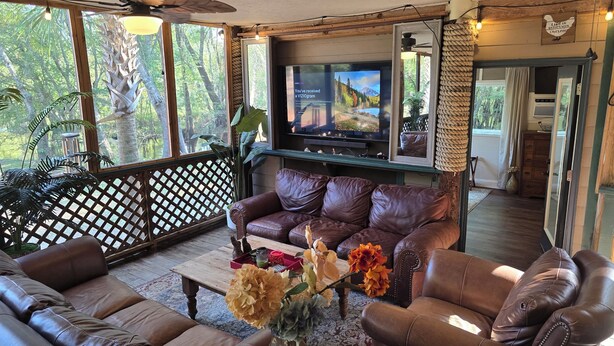 A spacious living room with leather sofas, a flat-screen TV, and a large window offering a view of trees. - River Front, Fishing, Kayaking, Boating, Tubing on the Edisto River   (Ridgeville)