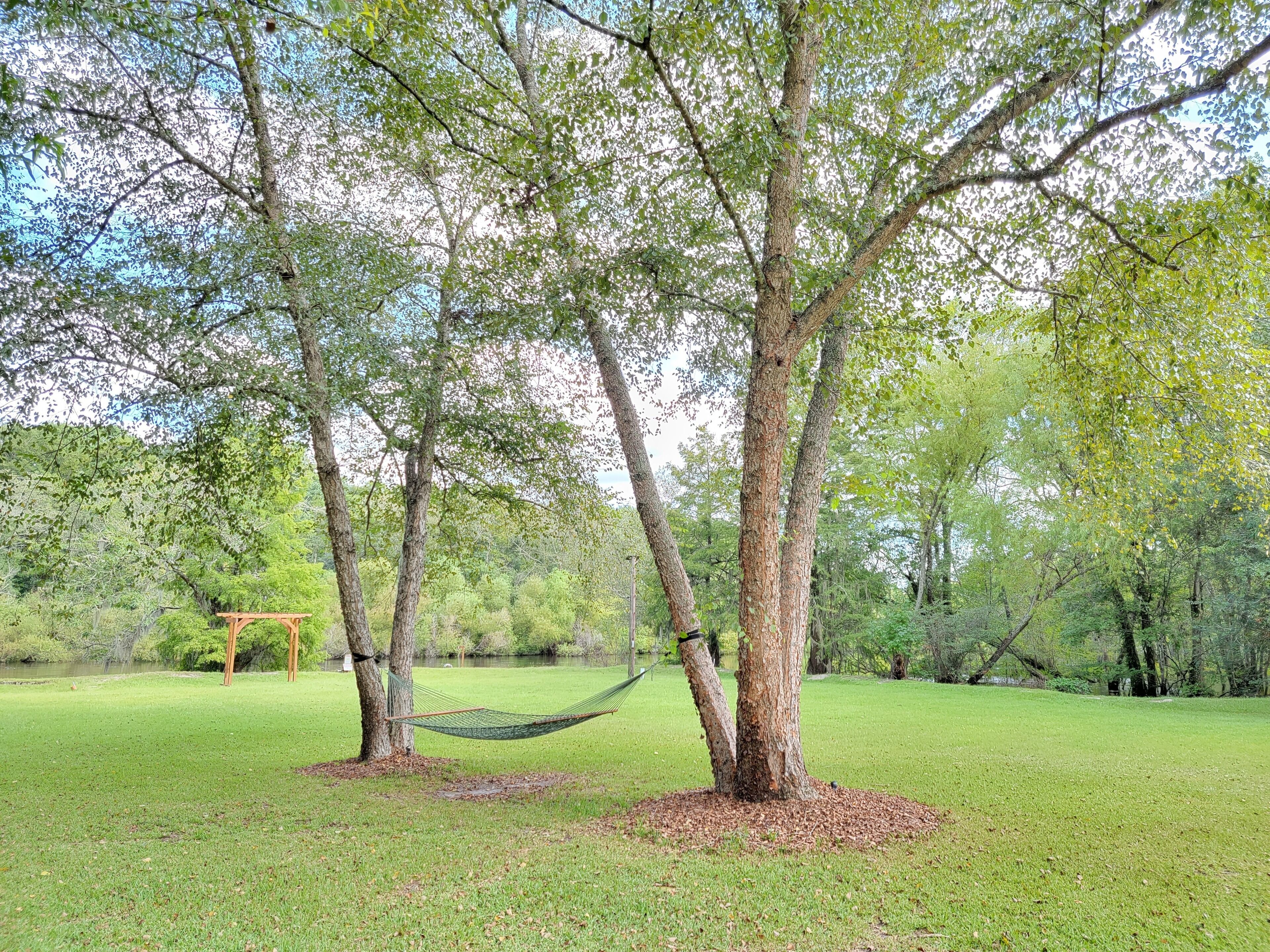 A hammock between two trees in a grassy park area with a wooden pergola in the background.