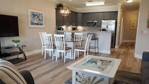 A modern living room with a sofa, a coffee table, and a TV. There is a dining area with chairs and a table. The kitchen is visible with cabinets and appliances. - Lakefront 311 - Updated Lake Front villa just steps to the beach and pools! (Miramar Beach)