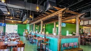 Ein Restaurant mit Holztischen und -stühlen, einer Theke mit grüner Wand und hängenden Pendelleuchten.