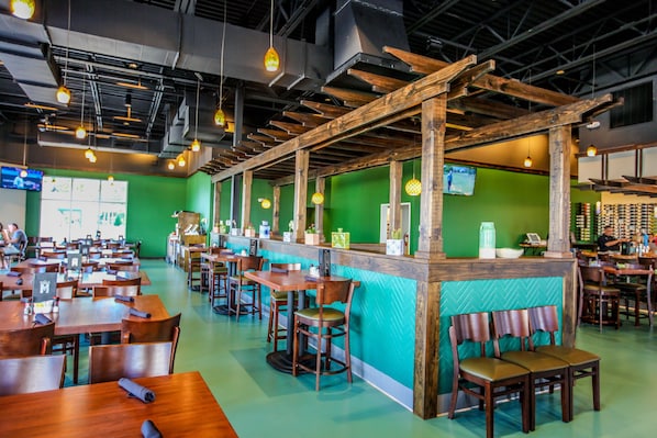 A restaurant with wooden tables and chairs, a counter with a green wall, and hanging pendant lights.