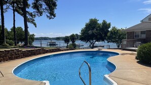 Una piscina con una escalera metálica, rodeada por un muro de piedra y un solárium de hormigón, con vistas a un lago y árboles al fondo.