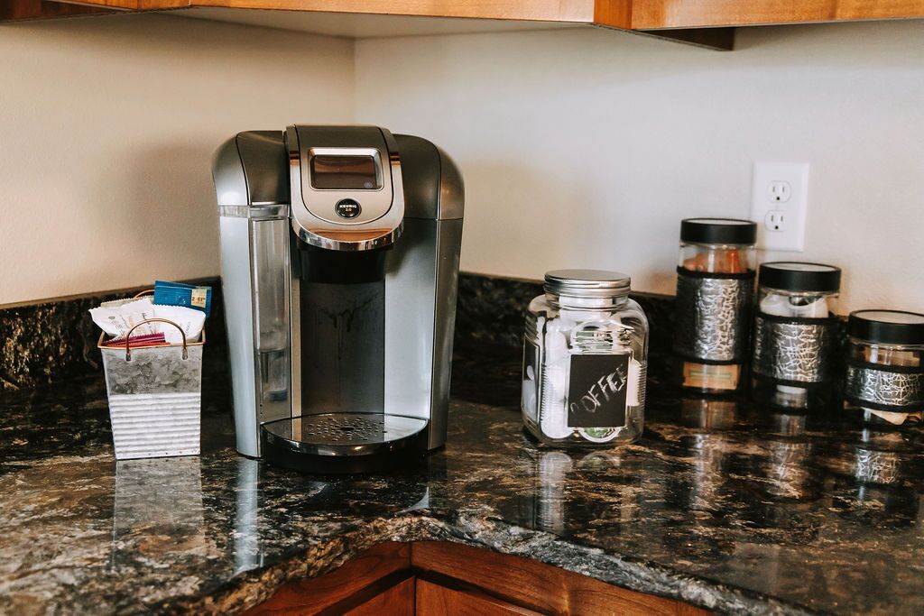 A coffee maker on a granite countertop with a basket of napkins and a jar of coffee pods.