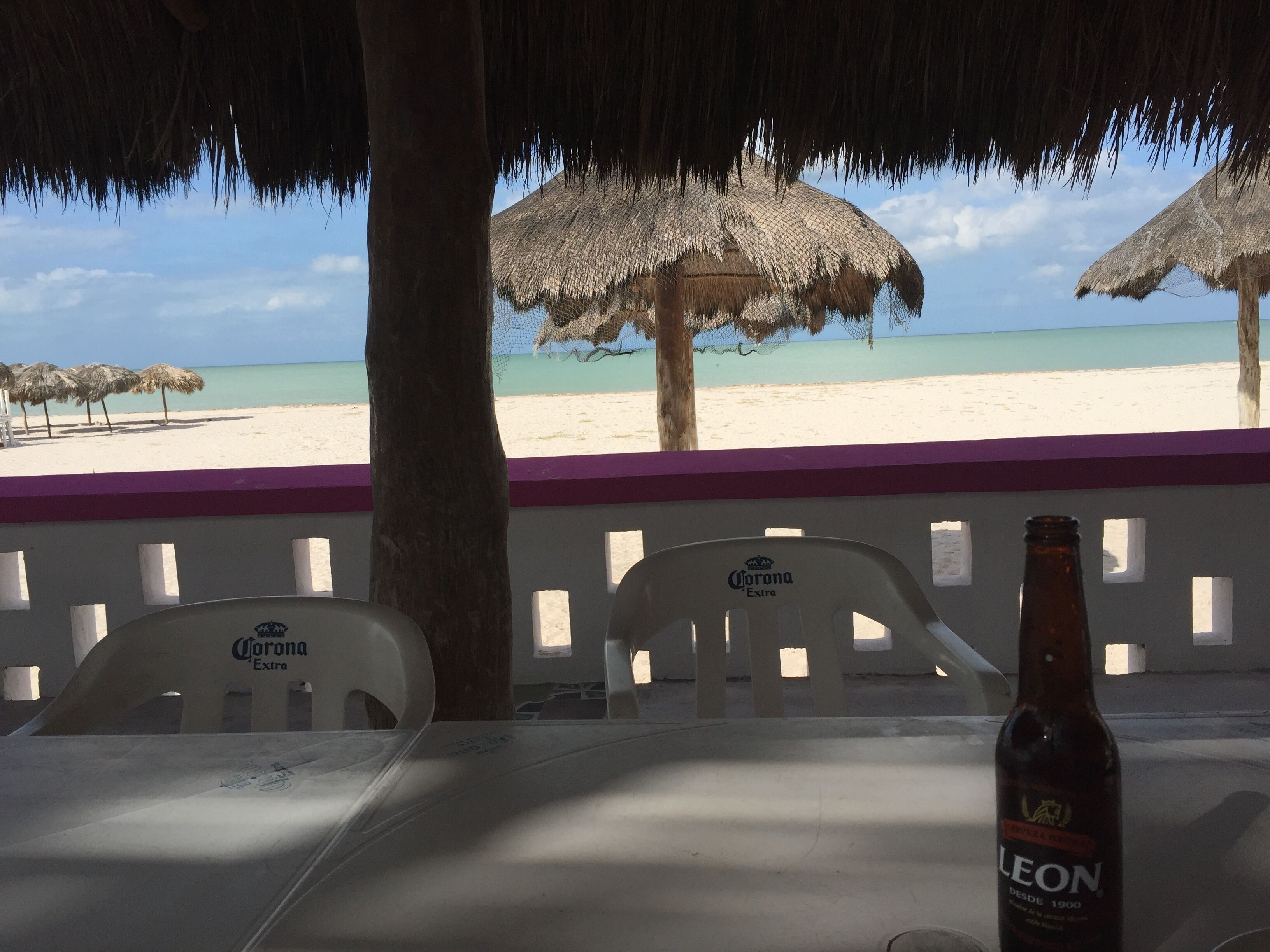 A beach view with a table, two chairs, a bottle of Leon beer, and thatched umbrellas.