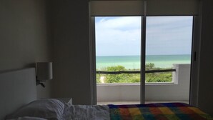Ein Hotelzimmer mit Bett, einer farbenfrohen Steudecke, einem weißen Kissen, einem Nachttisch und Blick auf das Meer durch eine Schiebetür.