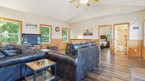 Un vaste salon comprenant un canapé en cuir, une table basse en bois et un téléviseur. On y trouve également un ventilateur de plafond en bois, de grandes fenêtres équipées de stores et un couloir menant à une salle de bain.