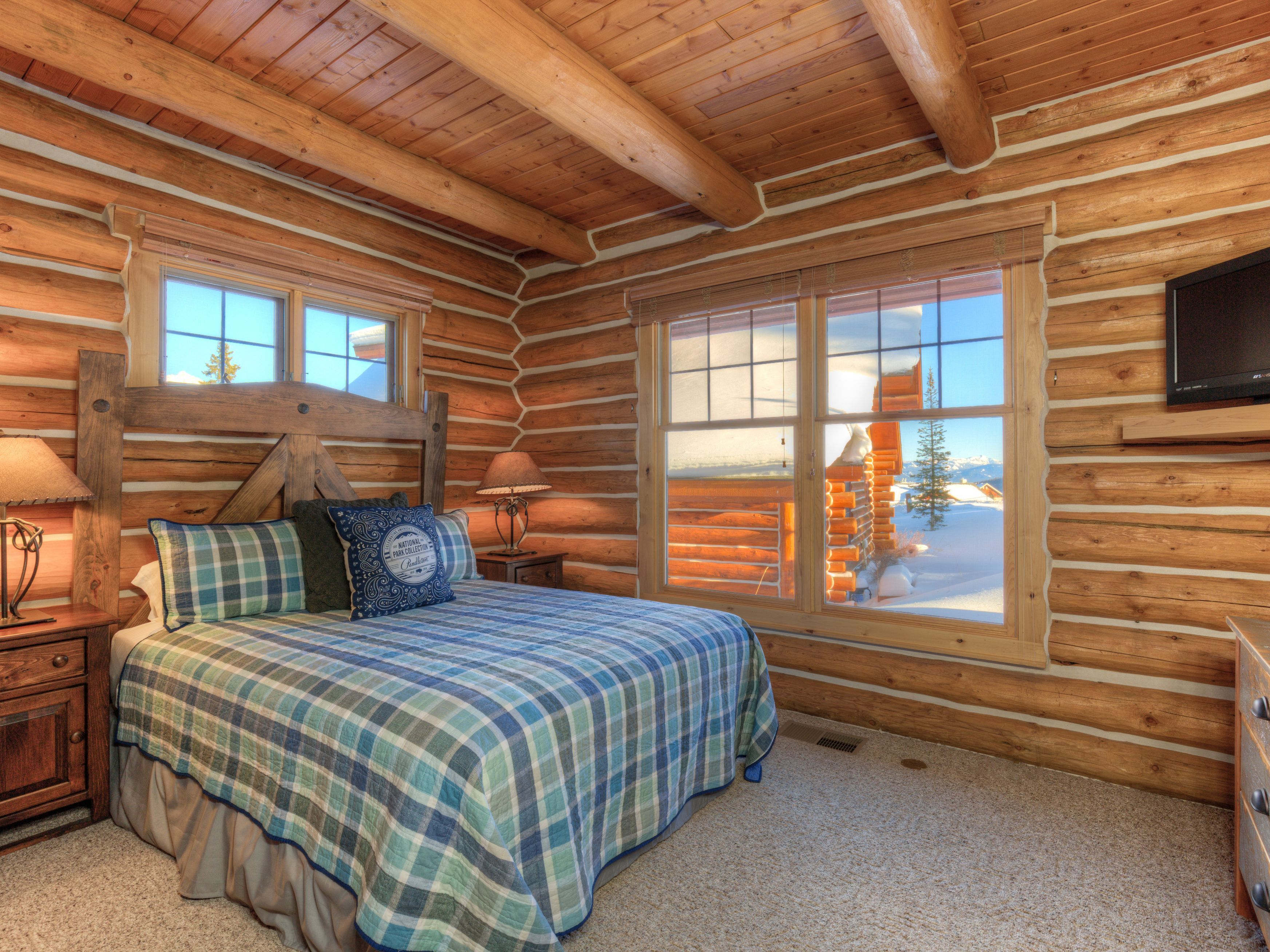 Ein Schlafzimmer in einer Blockhütte mit einem Bett, zwei Nachttischen, einem Fernseher und einem Fenster mit Blick auf Schnee.