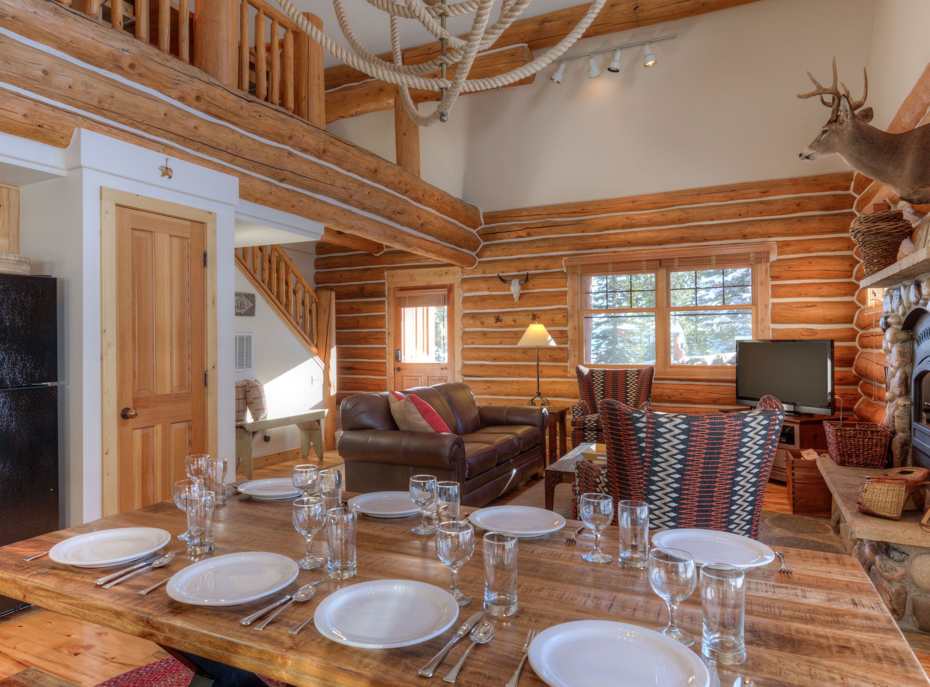 A log cabin interior with a dining area set for six, a fireplace, a mounted deer head, and a cozy living space with a sofa and TV.