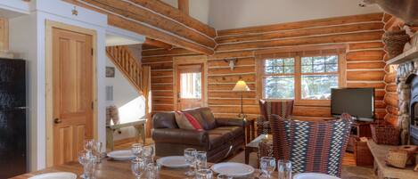 A log cabin interior with a dining area set for six, a fireplace, a mounted deer head, and a cozy living space with a sofa and TV.