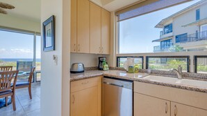 A modern kitchen with a dishwasher, a coffee maker, and a sink. There is a dining area with chairs and a table. The kitchen has large windows with a view of a building and greenery. - Kapalua Bay Villa 35G4 | Sunset Views | Walk to Beach (Lahaina)