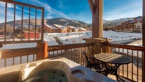 En træterrasse med jacuzzi og et bord, med udsigt over et sneklædt landskab med bygninger og bjerge.