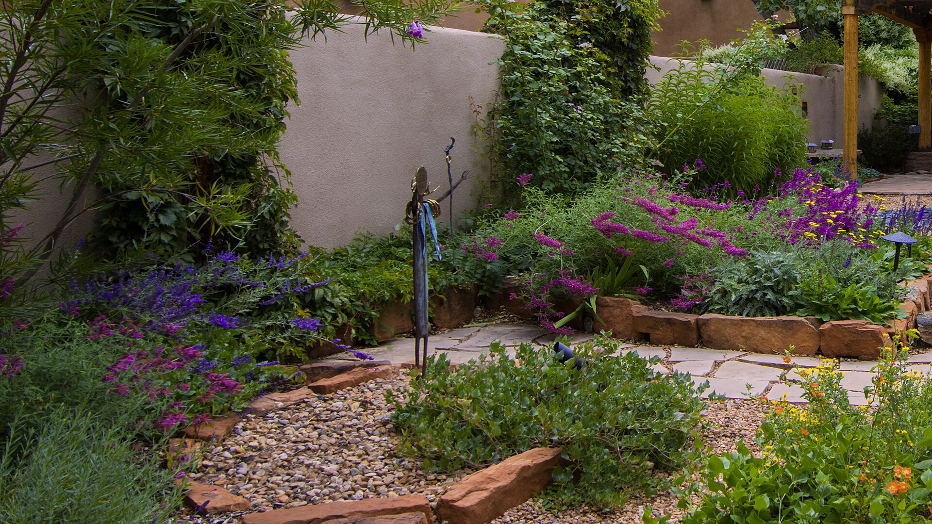 Un jardín con diversas plantas y flores, una pérgola de madera y un camino de piedra.