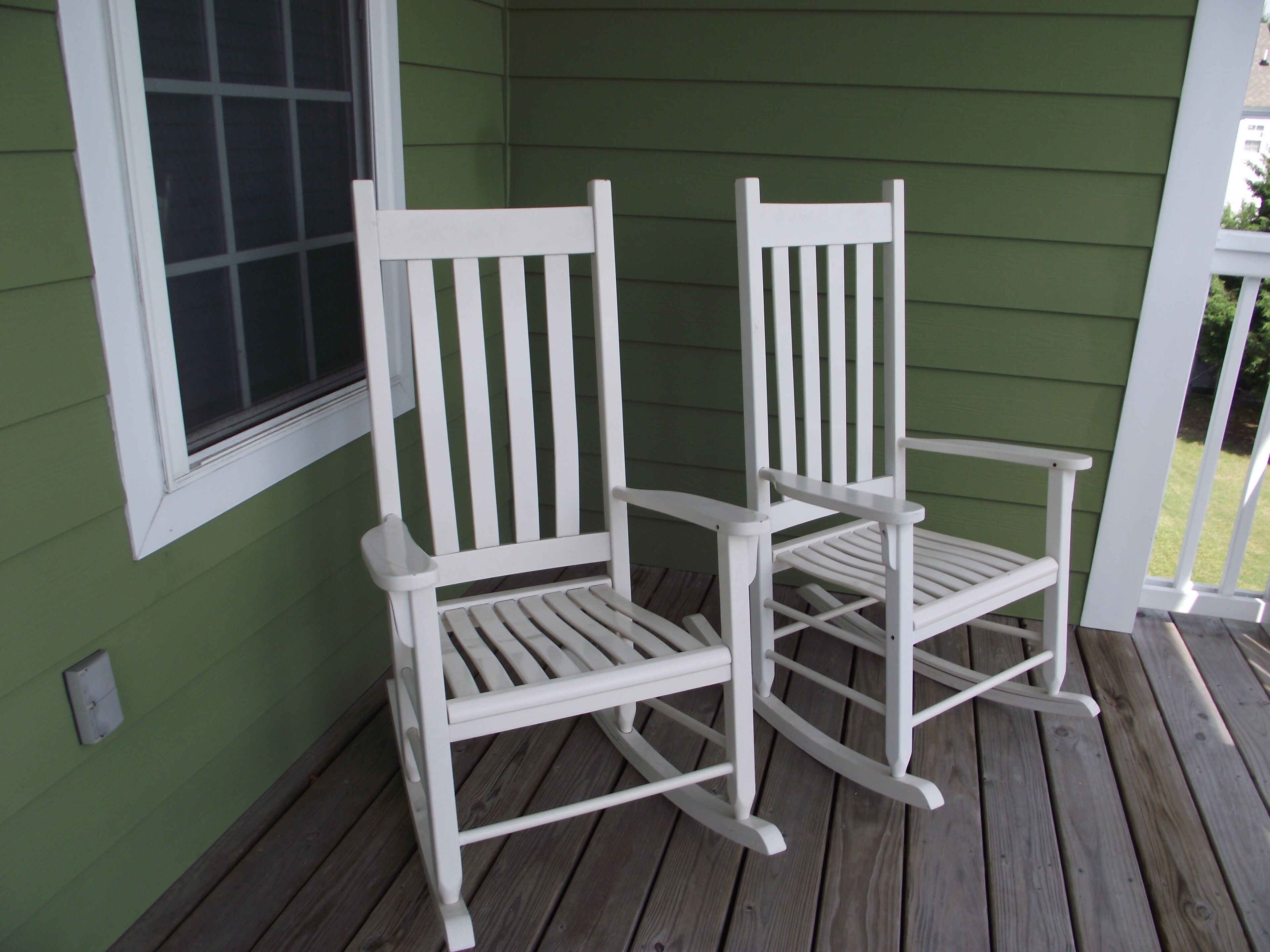 Two white rocking chairs on a wooden deck with a green wall and a window in the background.