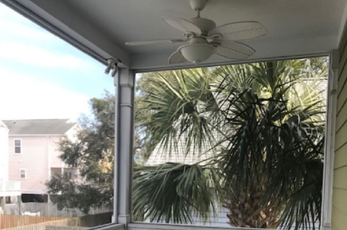 Ein Zimmer mit Deckenventilator, großem Fenster und Blick auf eine Palme sowie umliegende Häuser.