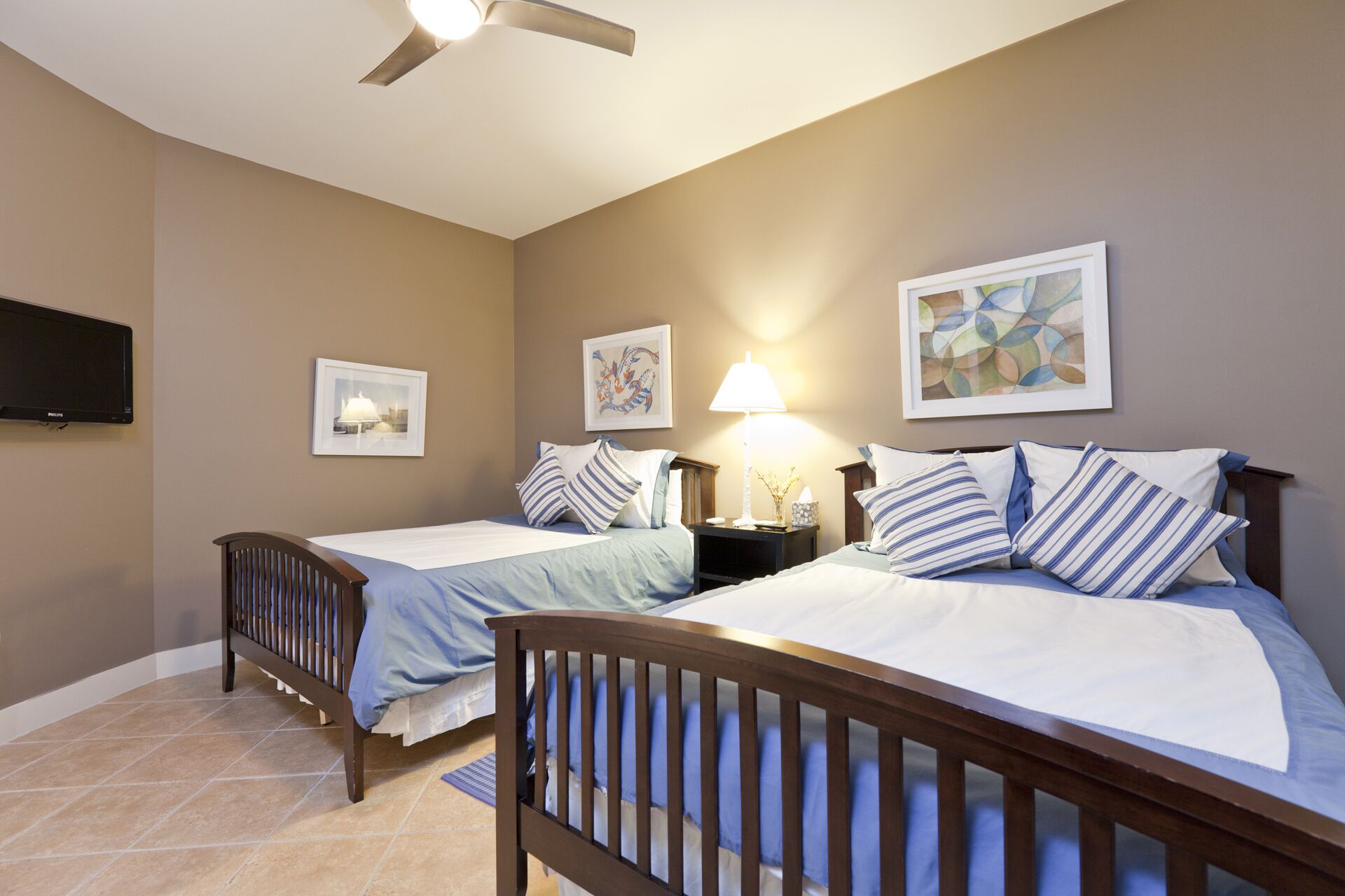 A bedroom with two beds, a ceiling fan, a flat-screen TV, and artwork on the walls.