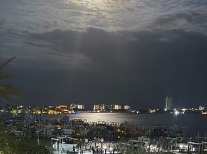 Una marina de nit amb vaixells amarrats, un perfil urbà al fons i un cel nuvolós.