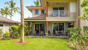 Una casa de dos pisos con porche cubierto, mobiliario exterior y un jardín bien cuidado con palmeras y arbustos.