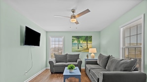 Un salon doté d’un ventilateur de plafond, d’un téléviseur à écran plat, de deux canapés gris, d’une table basse agrémentée d’une plante et d’un tableau représentant une scène de plage.