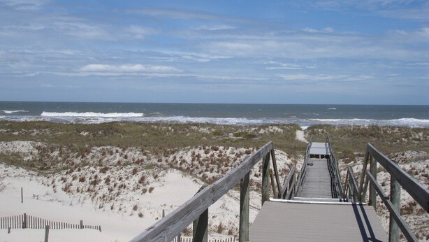 A wooden boardwalk leading to the beach with sandy dunes and the ocean in the background.