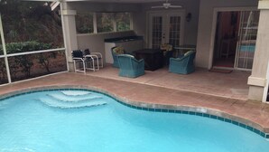 A swimming pool with a tiled edge, surrounded by a covered patio area with wicker furniture and a small kitchen area.