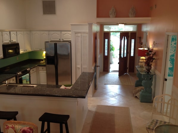 A kitchen with white cabinets, a stainless steel refrigerator, and a granite countertop. There is a wooden stool and a chair in the foreground.