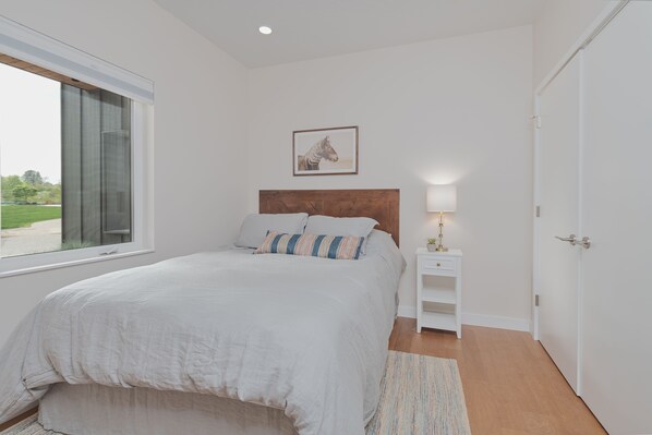 Une chambre à coucher avec un grand lit, une tête de lit en bois, une table de chevet avec une lampe, un tableau encadré au mur et une fenêtre donnant sur un paysage verdoyant.