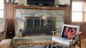 A living room with a stone fireplace, a television mounted above it, and a comfortable seating area with a chair and ottoman.