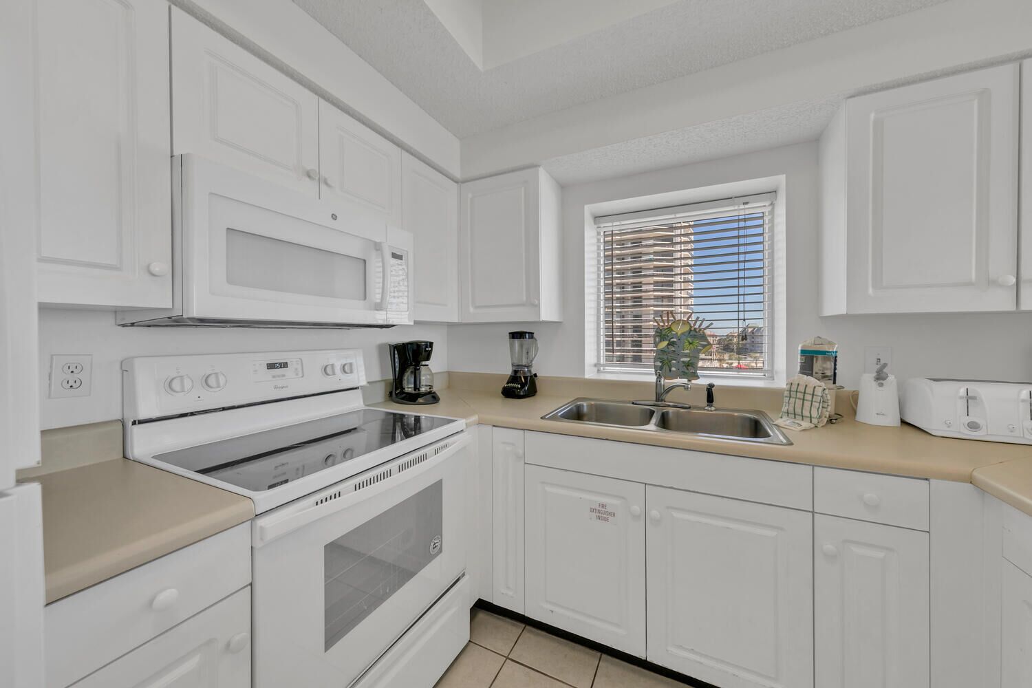 A modern kitchen with white cabinets, a stove, a microwave, a coffee maker, and a window with blinds.