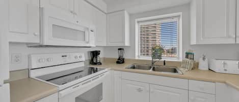 A modern kitchen with white cabinets, a stove, a microwave, a coffee maker, and a window with blinds.