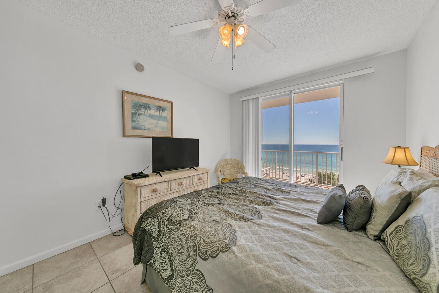 A bedroom with a bed, a television, a ceiling fan, a window with a view of the ocean, and a painting on the wall.