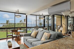 A modern living room with a large window overlooking a scenic view, a grey sofa with patterned cushions, and a dining area with chairs and a table. - 513 - 5th floor 1/1.5 Oceanfront unit with A/C and a great ocean view! (Kihei)