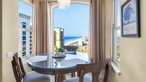 A dining area with a glass table, wooden chairs, and a view of the beach. - Beach Colony West 17F Penthouse/Gulf Front 3BR & Beach Service (Navarre)