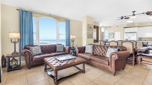 A spacious living room with a large window, a brown leather sofa set, a wooden coffee table, and a dining area with a kitchen in the background. - Beach Colony West 17F Penthouse/Gulf Front 3BR & Beach Service (Navarre)