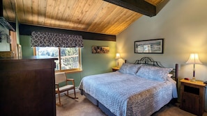 Une chambre avec un plafond en bois, un lit avec une literie à motifs, deux lampes de chevet, un fauteuil à bascule, un bureau et une fenêtre donnant sur des arbres.