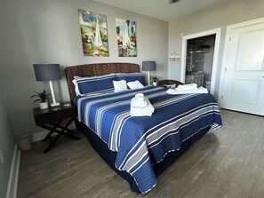 A neatly made bed with a striped blue and white bedspread, two wall-mounted paintings, a white door, and a small plant on a bedside table. - Top floor condo with amazing Gulf Coast view, steps away from! Pointe West Club (Galveston)