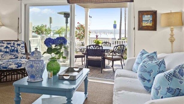 A living room with a white sofa, blue patterned cushions, a blue table with decorative items, and a view of the outdoors through large glass doors.