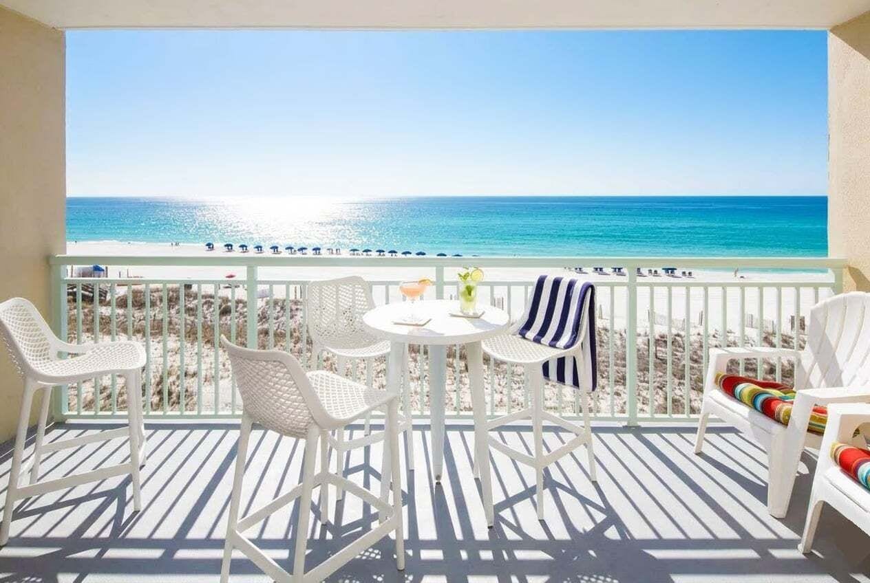 A balcony with a view of the beach, white wicker chairs, a small round table, and a striped towel.