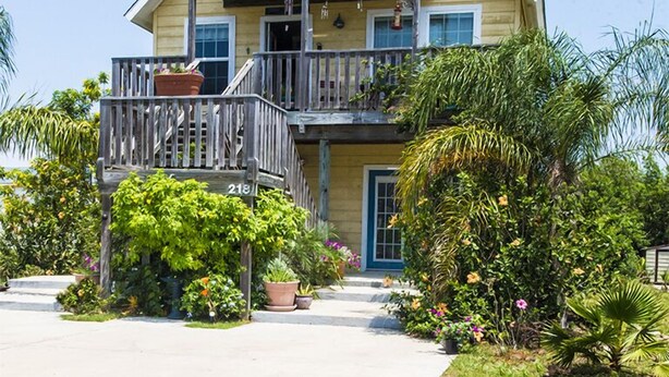 A two-story house with a balcony on the upper floor, surrounded by lush greenery and potted plants. - Island Serenity- Island Time Up in Old Town  (Port Aransas)