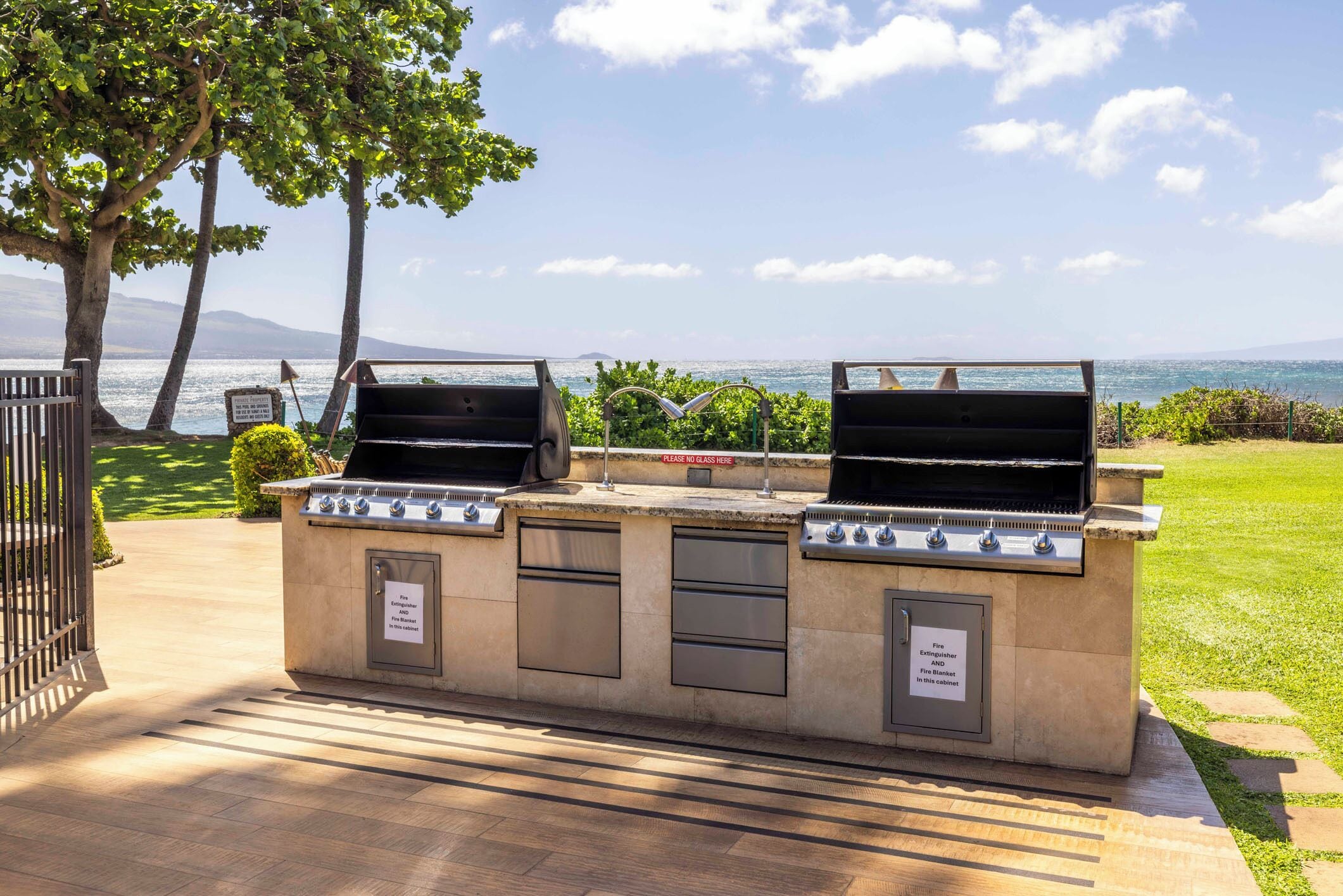 An outdoor grill station with a view of the sea, a fire extinguisher cabinet, and a 'Please no glass here' sign.