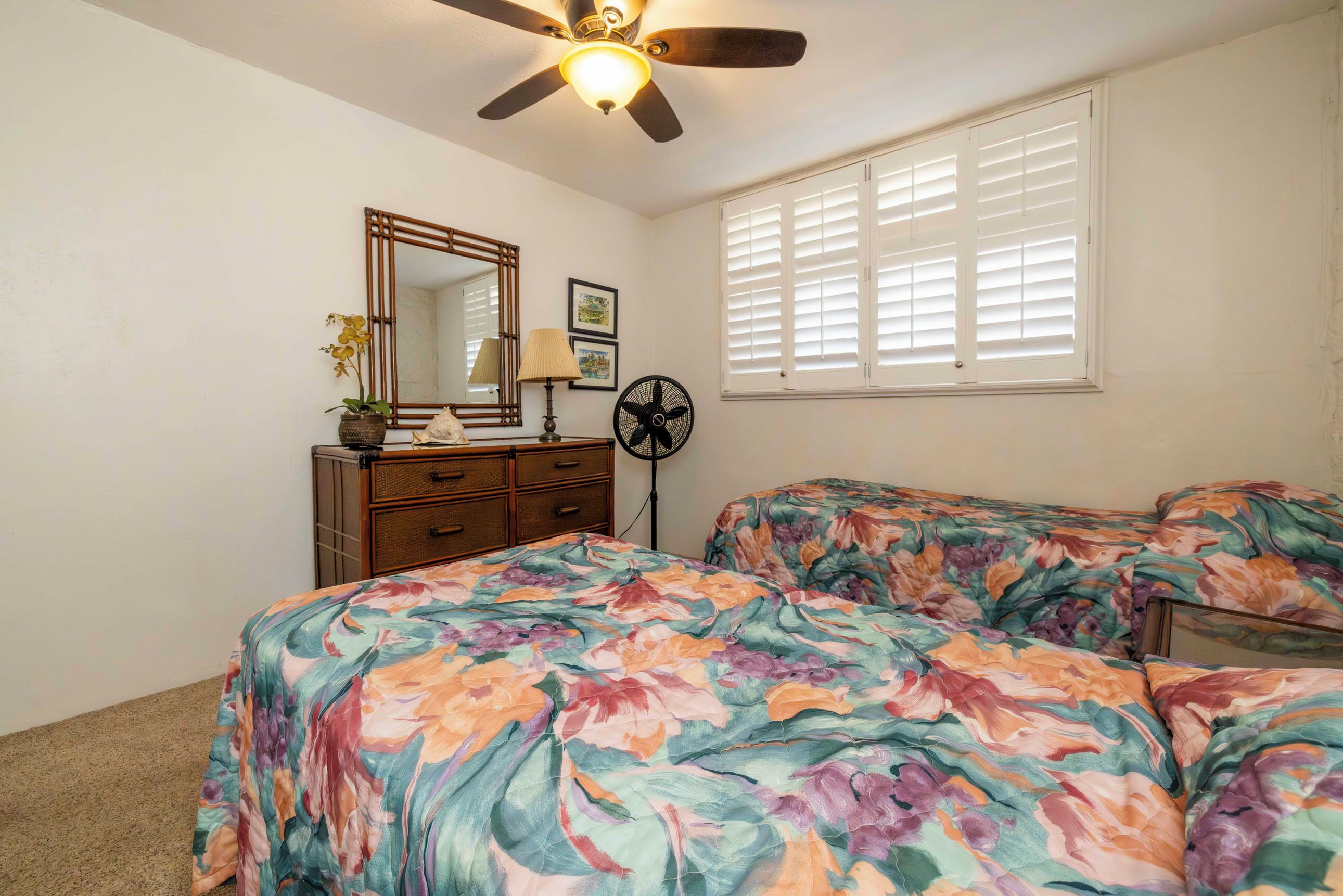 Une chambre à coucher comprenant un lit, une commode, un miroir, un ventilateur de plafond et une fenêtre avec des stores.