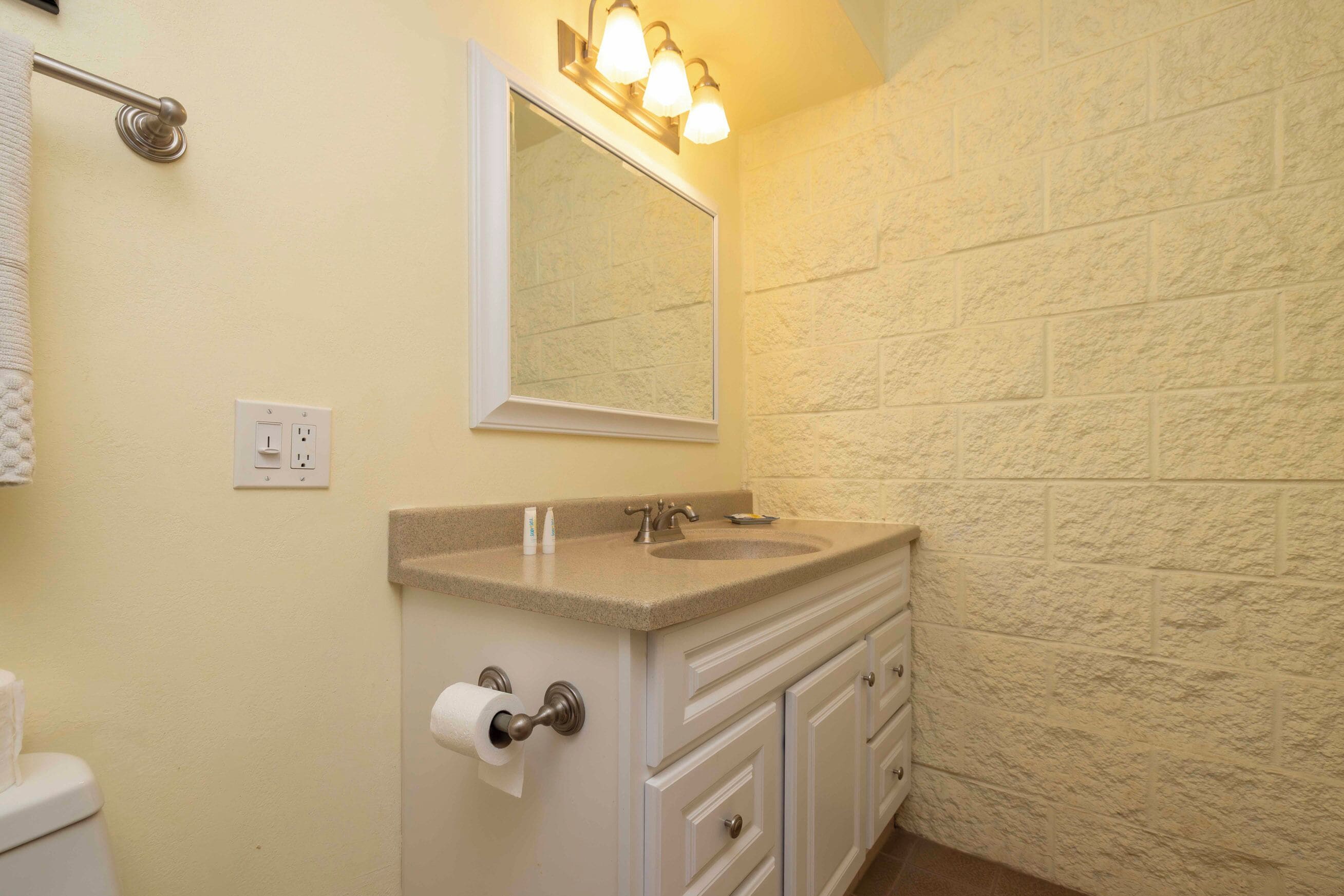 Un bagno con un piano di lavoro color beige, un lavandino, uno specchio, un porta asciugamani, un water e una lampada a parete.