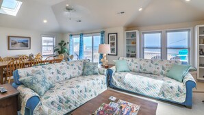 A living room with two patterned sofas, a coffee table with magazines, a dining area with wooden chairs, and large windows with a view. - 4759- Sun Daze - Ocean Views, Pool, Hot Tub & Steps to Beach (Kitty Hawk)