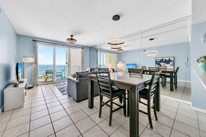 Amplia sala de estar con mesa de comedor, sillas y sofá. Cuenta con ventanas grandes con cortinas, un balcón y vistas al mar.
