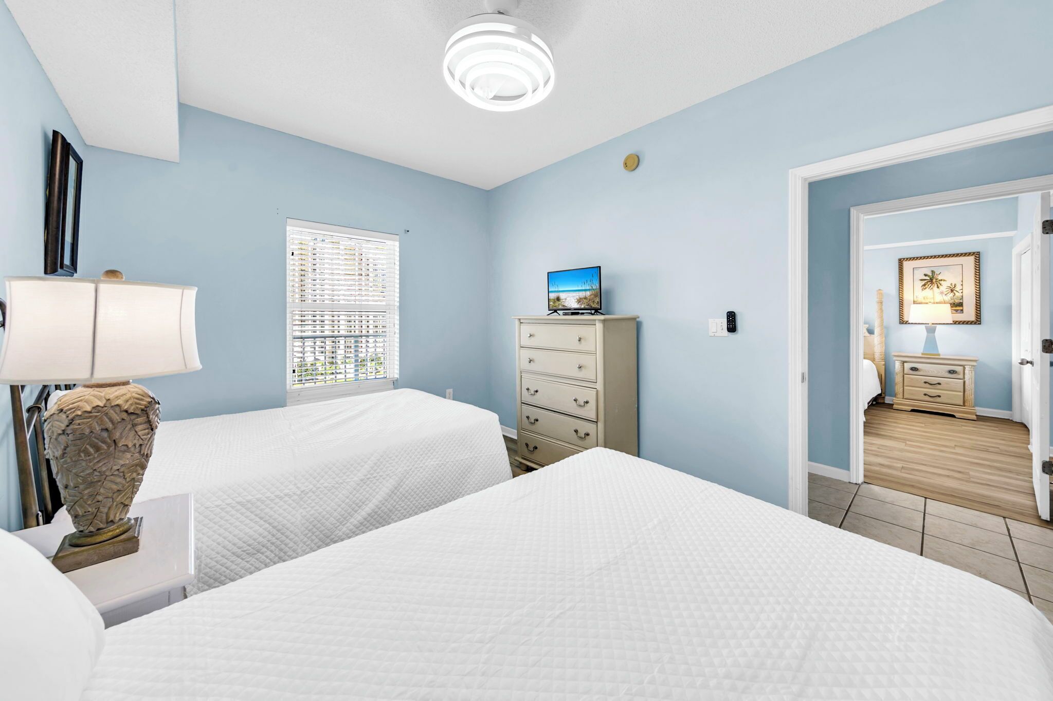 Een hotelkamer met twee bedden, een dressoir, een lamp, een televisie en een deur naar een andere kamer.