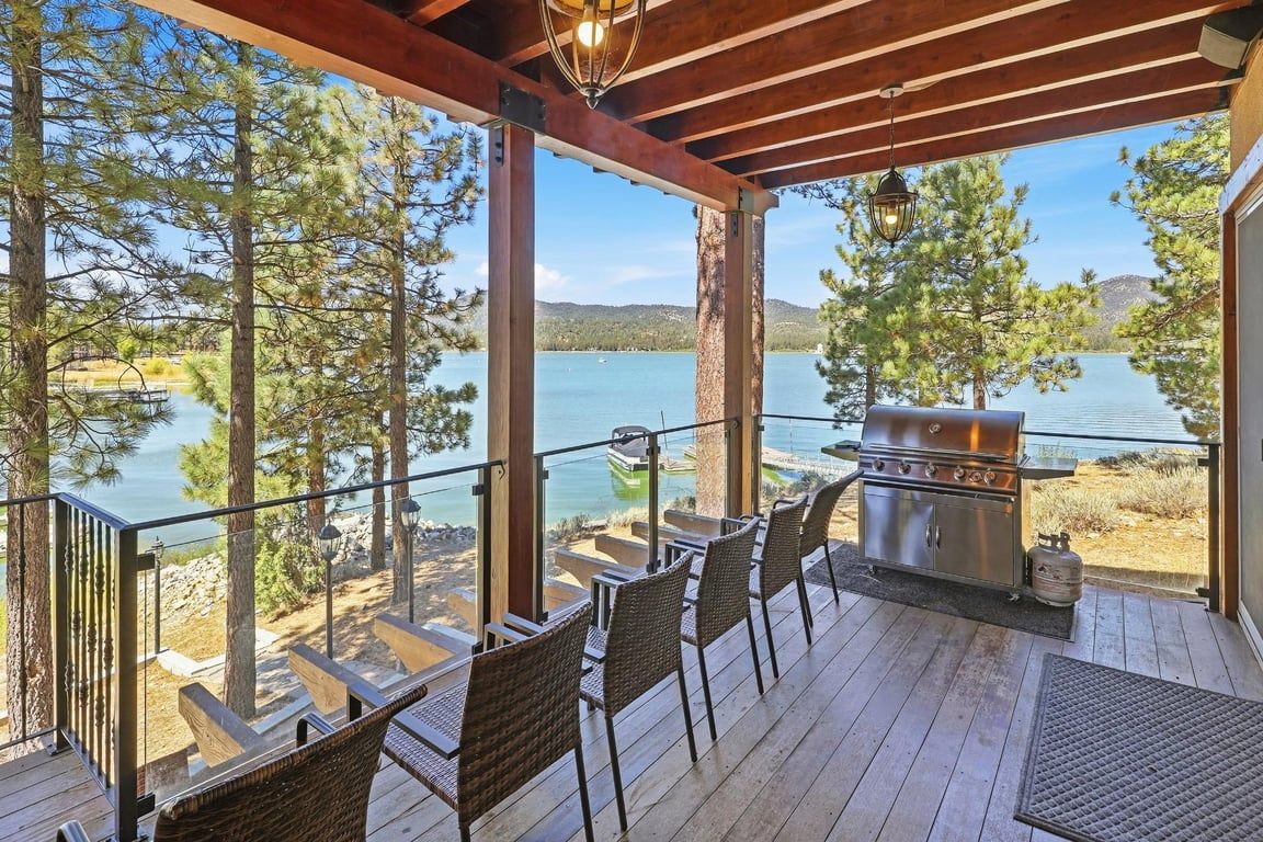 A covered deck with outdoor seating and a grill, overlooking a lake with trees and a distant mountain.