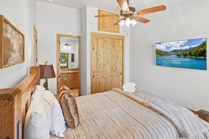 Een slaapkamer met een bed, een plafondventilator, een houten deur, een spiegel, een televisie en een ingelijst schilderij aan de muur.