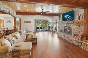 A spacious living room with a stone fireplace, wooden flooring, and a ceiling fan.