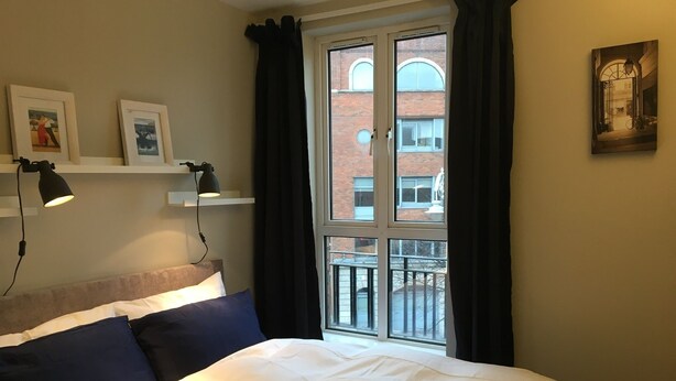 Un letto rifatto con biancheria bianca e blu navy, una testiera e due lampade da comodino. Una finestra con tende nere mostra un edificio in mattoni all'esterno.